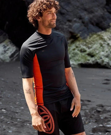 Man wearing black and orange short-sleeve rash vest and black shorts with red design on rocky beach