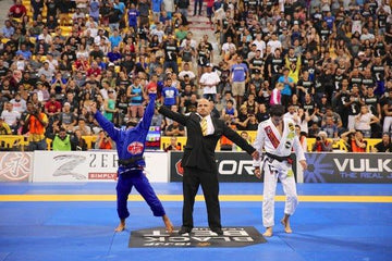 Brazilian Jiu-Jitsu match with referee raising winning competitor's hand in blue gi at crowded indoor tournament