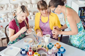 Three women painting colorful ceramic cups and pitcher in bright art studio with clay pottery shelves