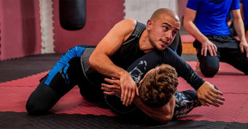 Two men grappling on red and black mats during Brazilian Jiu-Jitsu training in a gym setting