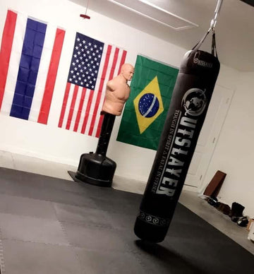 Indoor martial arts gym with punching bag, training dummy, and flags of Thailand, USA, and Brazil