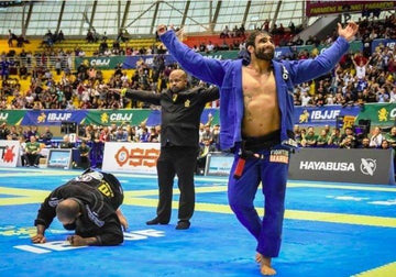 Brazilian Jiu-Jitsu athlete in blue gi celebrates victory on mat while opponent in black gi kneels and referee signals outcome at IBJJF tournament
