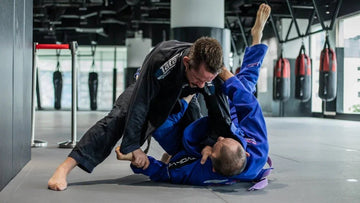 Two men sparring in Brazilian Jiu-Jitsu class wearing gis in a modern gym with punching bags