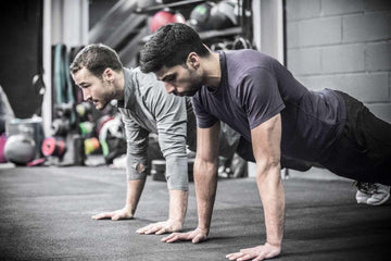 Two men doing pushups side by side in a gym with fitness equipment in background