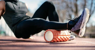 Person in black athletic wear using orange foam roller for leg muscle recovery outdoors on track
