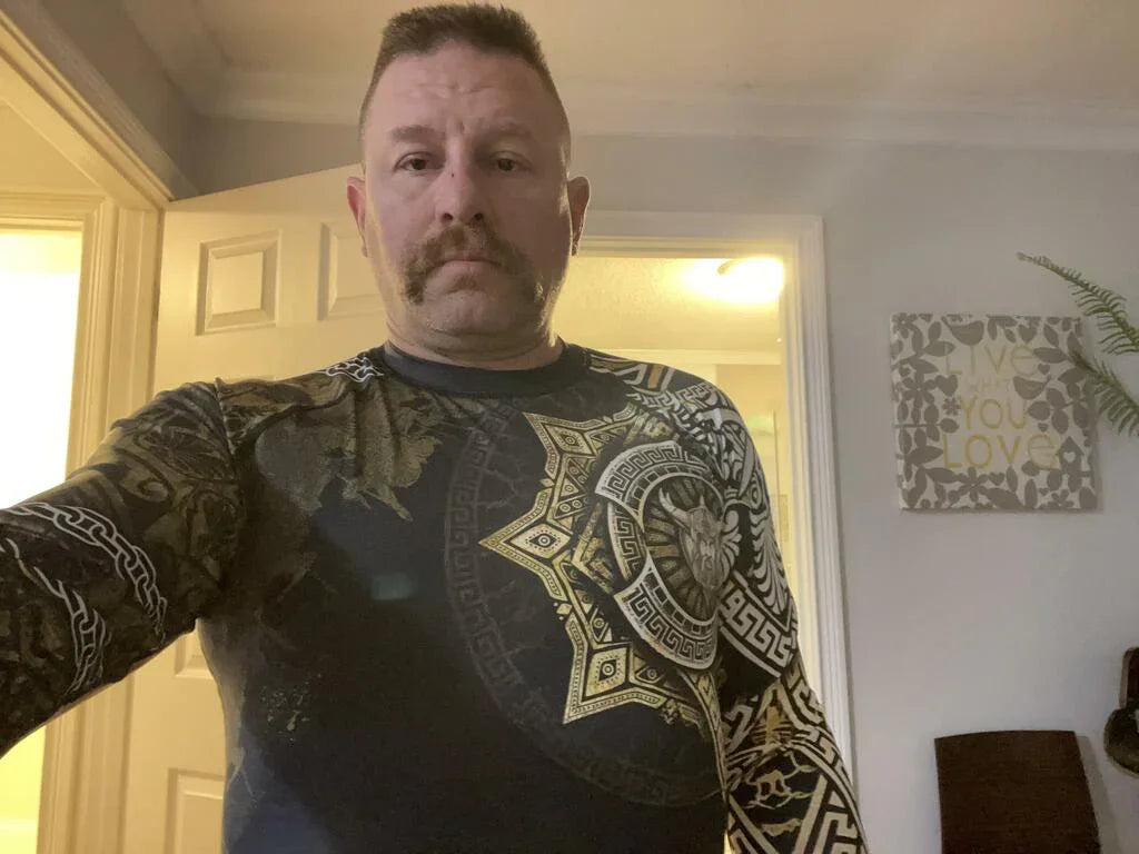 Man wearing black and gold Viking warrior medal long sleeve rash guard with intricate Norse designs indoors